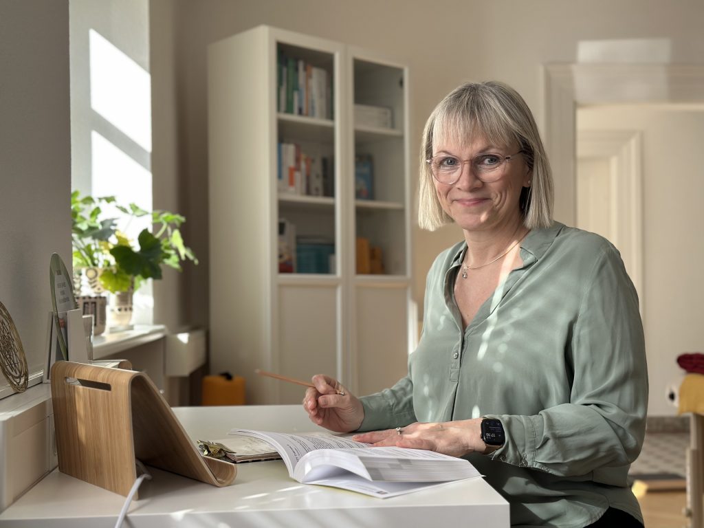 Heilpraktikerin mit blonden, schulterlangen Haaren sitzt an einem Tisch und studiert Unterlagen. Sie trägt eine hellgrüne Bluse und hält einen Bleistift. Im Hintergrund sind Bücherregale und Pflanzen sichtbar in der Praxis im Dreiländereck.
