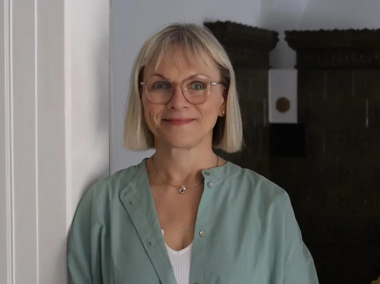 Heilpraktikerin mit blonden Haaren und Brille in grüner Bluse lächelt freundlich im Dreiländereck. Sie steht an einer Wand mit dunklem Hintergrund.