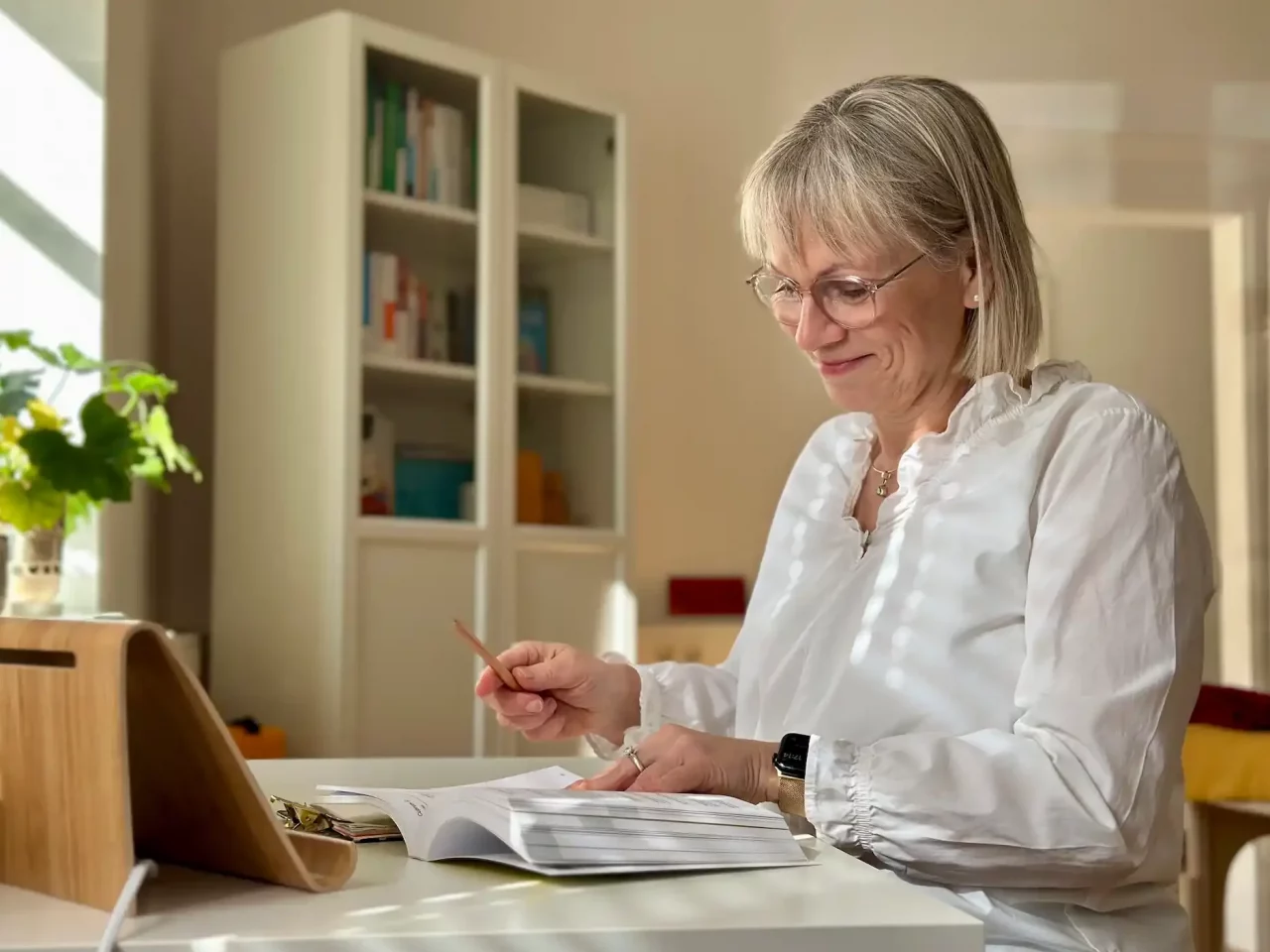 Blonde Katrin Zähle Heilpraktikerin trägt eine weiße Bluse und sitzt an einem Tisch. Sie hält einen Bleistift in der Hand und blättert in einem Buch. Im Hintergrund sind Regale mit Büchern und Pflanzen sichtbar.