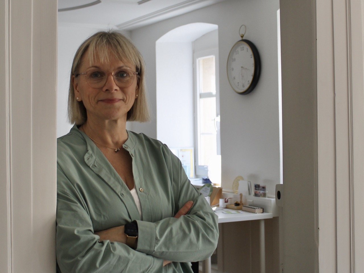 Blonde Katrin Zähle Heilpraktikerin mit schulterlangen Haaren, trägt eine grüne Bluse, steht mit verschränkten Armen im Türrahmen. Im Hintergrund sind ein weißer Tisch und eine Wanduhr sichtbar.