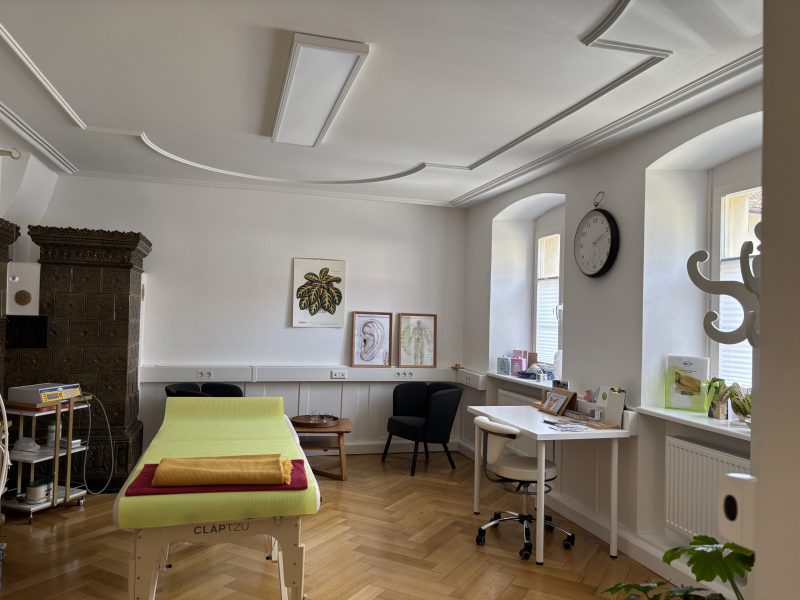 Ein Behandlungsraum im Dreiländereck mit einem grünen Behandlungstisch, einem schwarzen Stuhl, einem weißen Schreibtisch mit einem weißen Stuhl und mehreren Bildern an der Wand. Eine große Uhr zeigt die Zeit an.