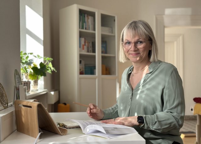 Heilpraktikerin mit blonden, schulterlangen Haaren sitzt an einem Tisch und studiert Unterlagen. Sie trägt eine hellgrüne Bluse und hält einen Bleistift. Im Hintergrund sind Bücherregale und Pflanzen sichtbar in der Praxis im Dreiländereck.